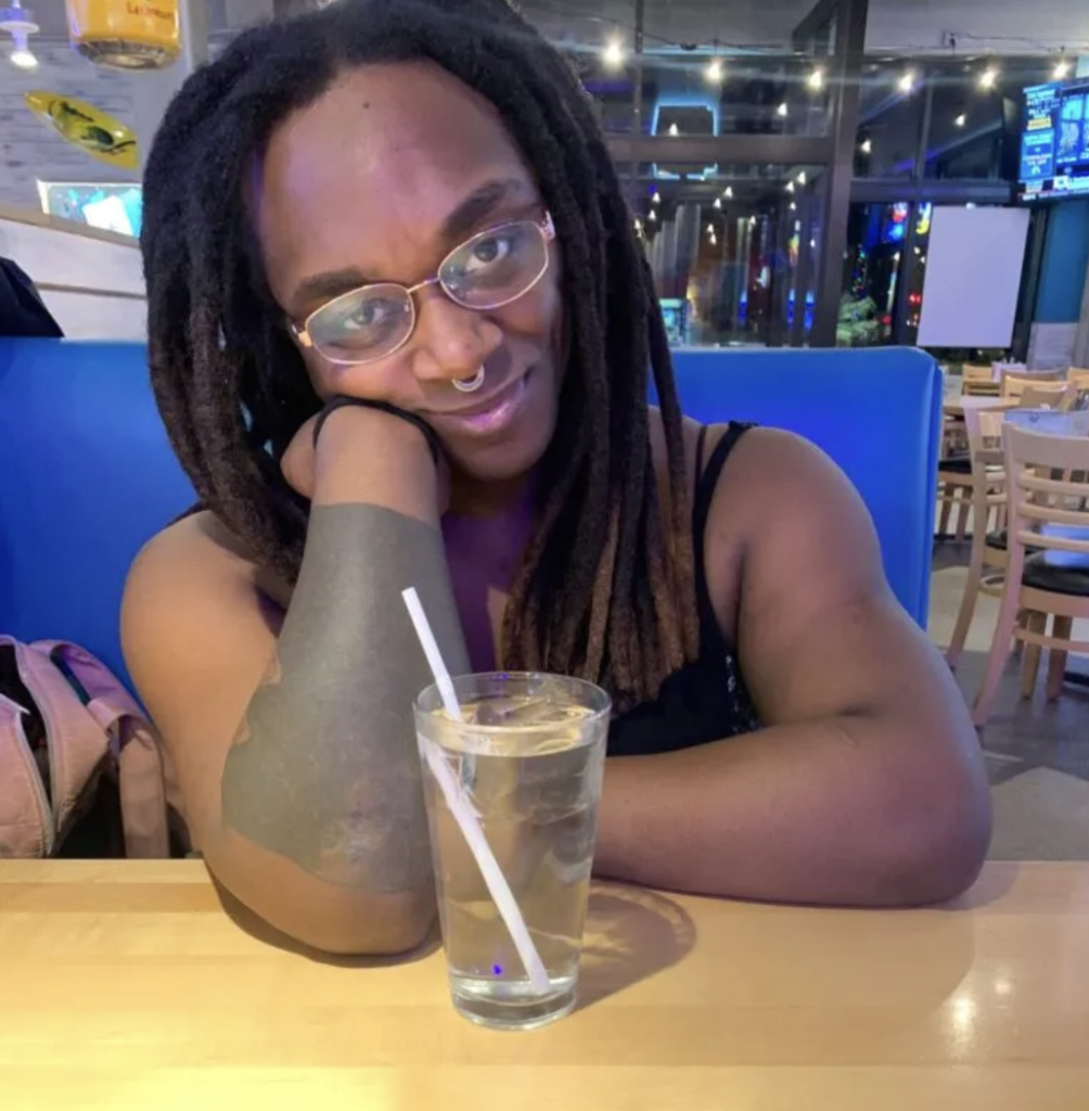 Person with long locs and glasses, resting their head on their hand at a restaurant table with a glass of water in front of them, smiling softly.